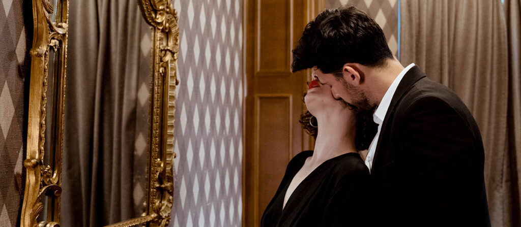 man and woman in front of a mirror, with the man sensually kissing the woman's neck.